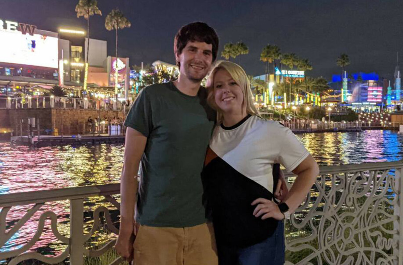 Smiling couple posing by a waterfront at night with colorful city lights in the background.